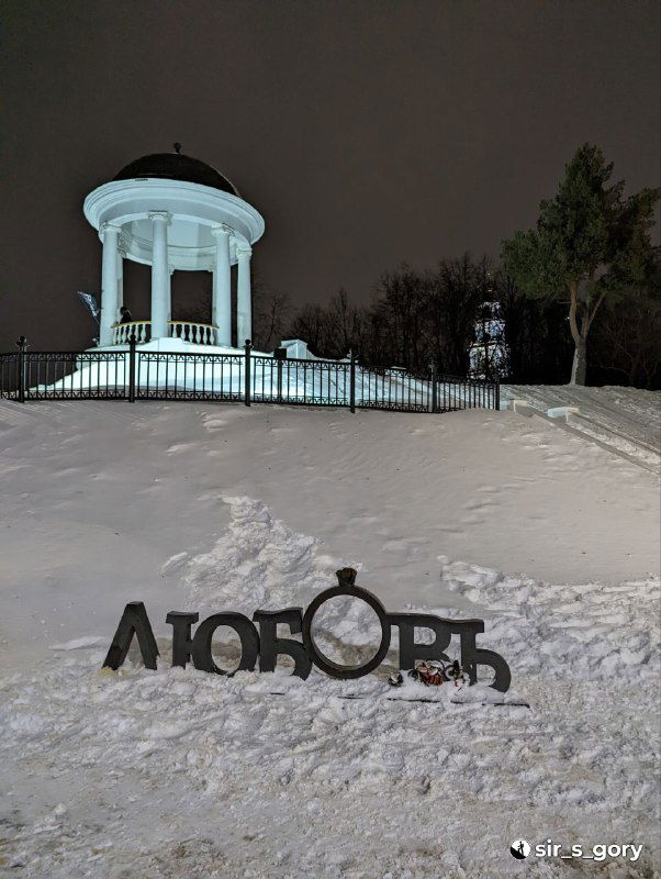 Заснеженная беседка Островского на холме вечером, белая ротонда и арт‑объект «Любовь» на переднем плане в городской зимней сцене.