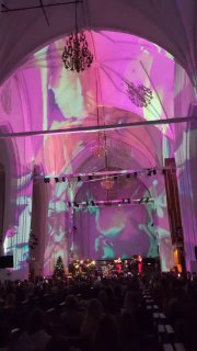 Интерьер Кафедрального собора с разноцветными проекциями и сценой; видно публику и музыкальную группу, создающую рок-версию классических тем.