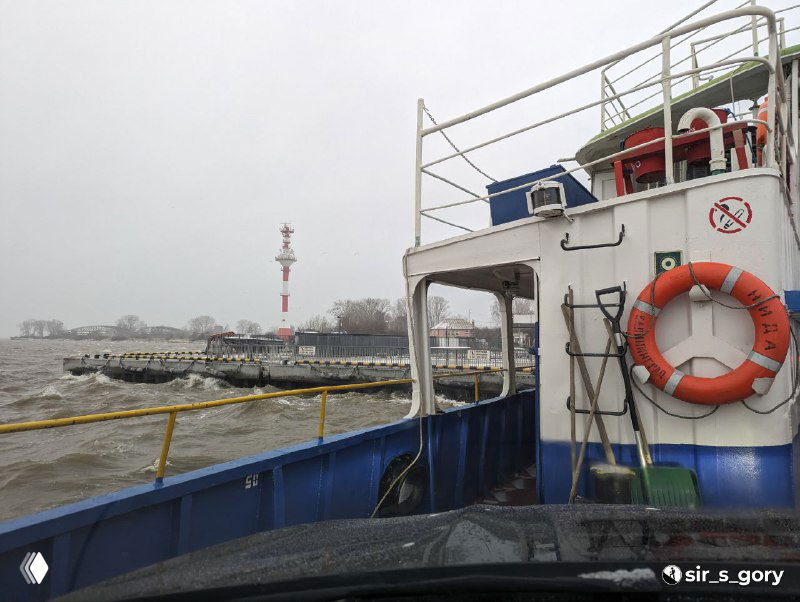 Вид с парома на вход в мол и удаляющийся горизонт: белесая вода, береговая линия и редкие строения на косе в морозный день