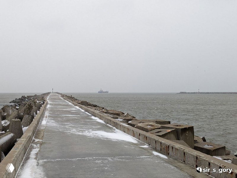 Длинный бетонный мол, устремленный в море: поверхность покрыта тонким слоем снега, по краям видны волны и туманные силуэты вдалеке