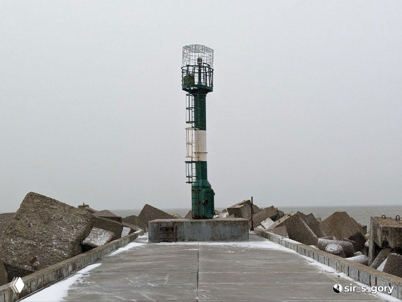 Ближайший план зеленого маяка на конце мола: покрытая снегом дорожка, бетонные блоки и тихая Балтика за маяком в пасмурный день