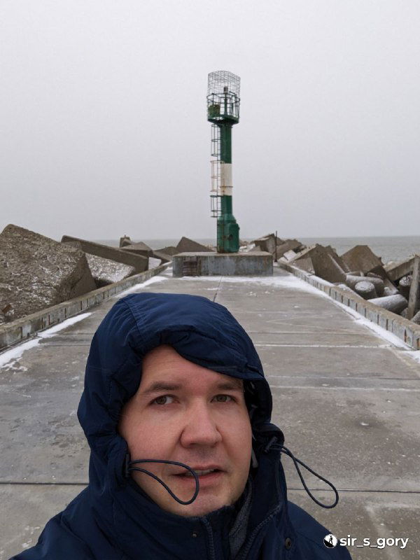 Селфи автора на молу с зеленым маяком позади: лицо в капюшоне, ветровка, снежные брызги и ощущение холода у моря