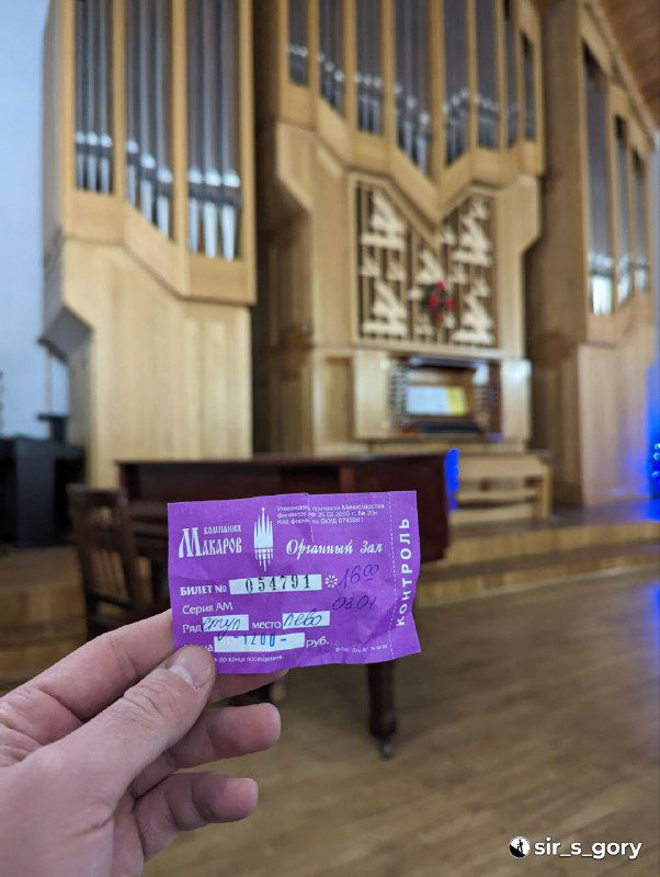 Билет на концерт в органном зале на переднем плане, в глубине виден интерьер зала и трубы органа — кадровый ракурс с рукой держателя билета.