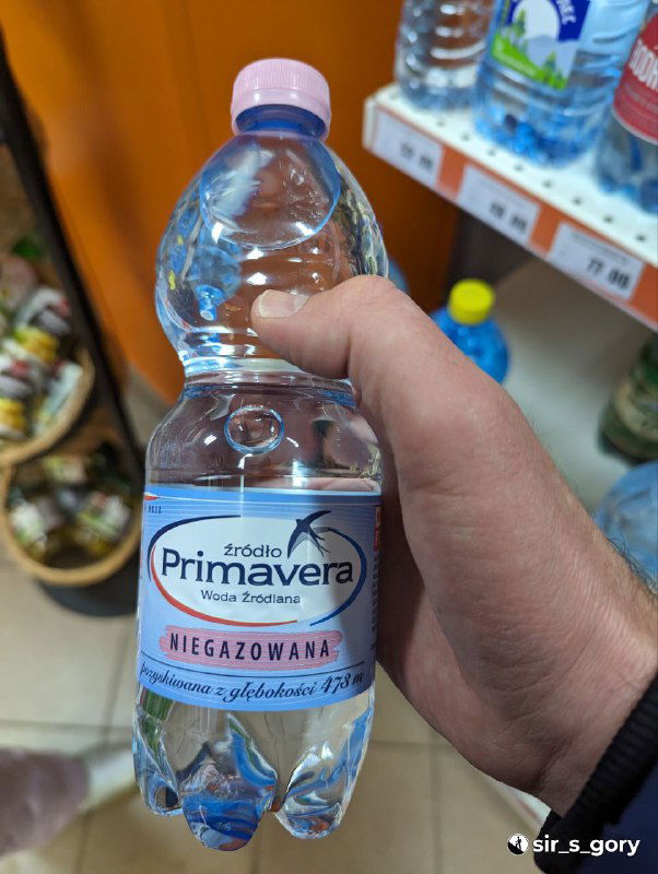 Рука держит бутылку польской воды Primavera 1 л в магазине: выпуклая этикетка на пластиковой таре и полки с товарами на фоне.