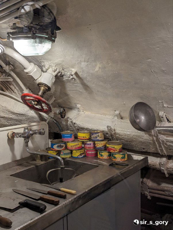 Камбуз подводной лодки: мойка, стопка консервных банок и кухонные принадлежности в тесном помещении, иллюстрирующие быт экипажа на борту.