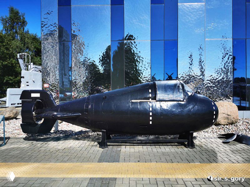 Сверхмалая подводная лодка «Тритон-1М» на открытой площадке музея Мирового океана: маленький корпус на постаменте, летняя экспозиция на фоне зелени.