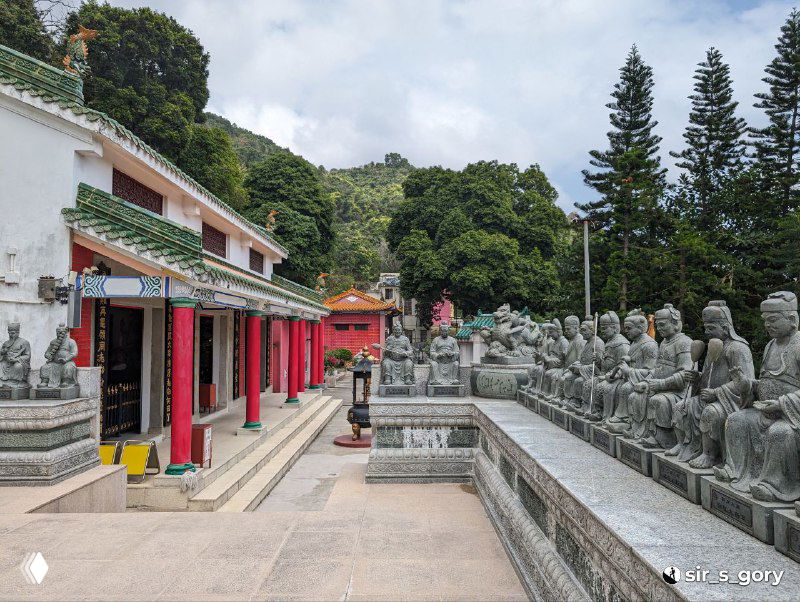 Ряд каменных и позолоченных скульптур вдоль дорожки в монастырском дворе, видно постройки комплекса и зелёные холмы вокруг.