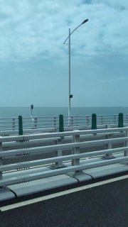 Кадр с моста: перила и опора освещения на фоне моря, вид на полосу движения и открытый морской простор в ясный день с легкой облачностью.