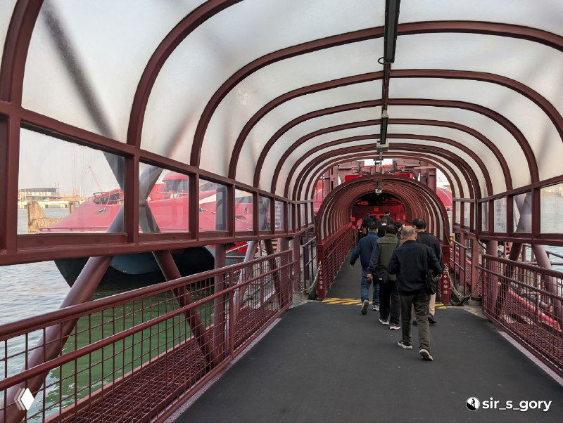 Покрытый пешеходный переход с красной арочной металлической конструкцией, люди идут по коридору над водой — подход к причалу или переход между зонами.