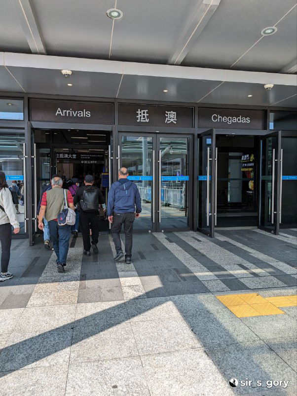 Вход в зону прибытия Макао: автоматические двери с надписями Arrivals и Chegadas, пассажиры с багажом проходят внутрь терминала по выделенной дорожке.