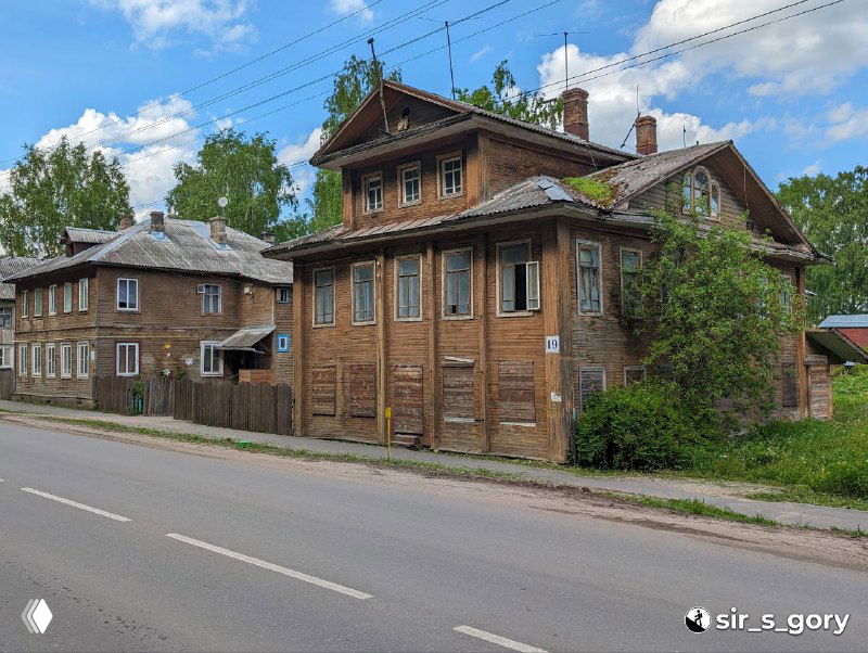 Ряд старых купеческих деревянных домов вдоль улицы с традиционной резьбой и выцветшими фасадами, улица без активной торговли летом.