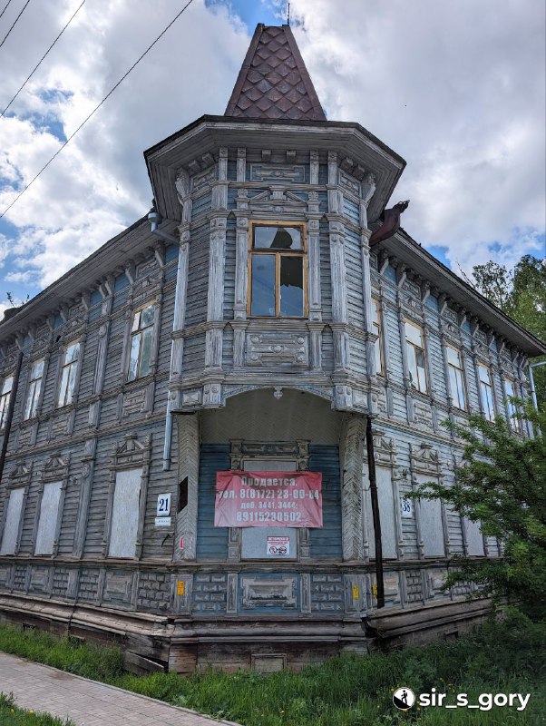Угловой вид на деревянный особняк с вывеской «Продам» на фасаде, трещины и следы старения на декоративных элементах здания.