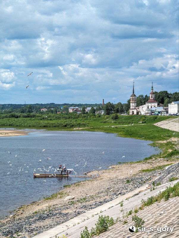 Пейзажный кадр реки с прибрежной косой, корабликом и стайкой чаек; на дальнем берегу видны церковные силуэты и зелёные склоны.