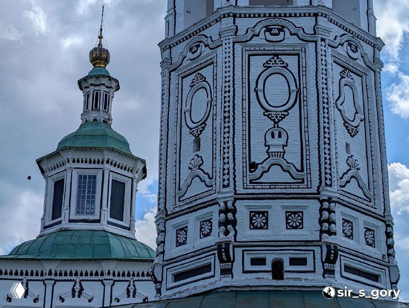 Крупный фрагмент расписного фасада церковной стены с росписями и орнаментом на фоне голубого неба; архитектурные декоративные детали.