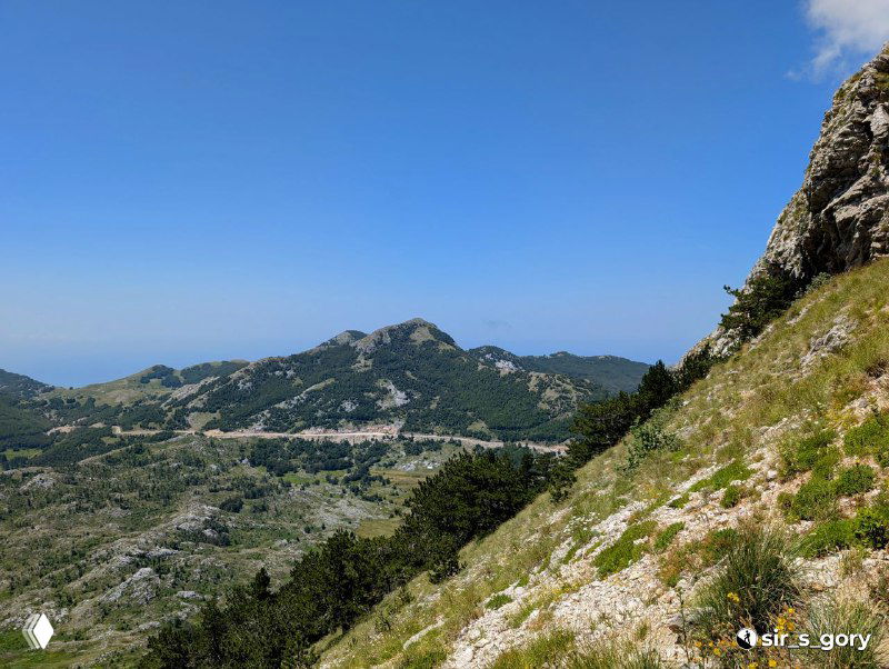 Широкий вид с горного склона на расчленённый рельеф и дальние горы, вдалеке виднеется полоса моря на горизонте под голубым небом.