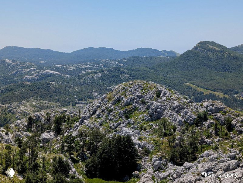 Каменистые склоны и заросли низкорослой растительности на переднем плане, далее — гряды гор с мягкими очертаниями на фоне неба.