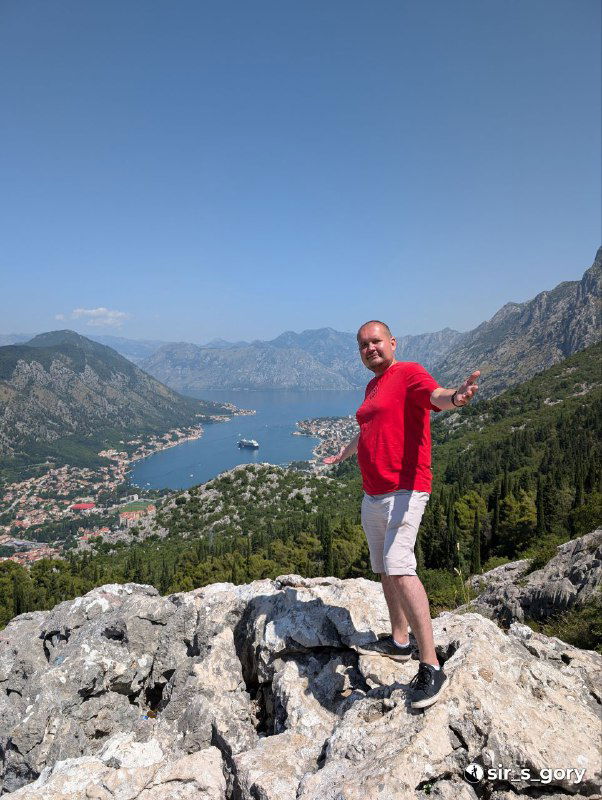 Турист в красной футболке стоит на каменной осыпи на фоне Которского залива — фигурный план на фоне панорамного вида.