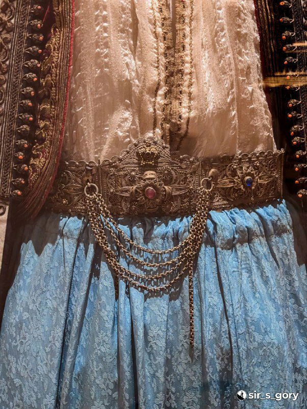 Крупный план серебристого пояса с декоративными цепочками и фактурной тканью юбки традиционного черногорского костюма в витрине музея.