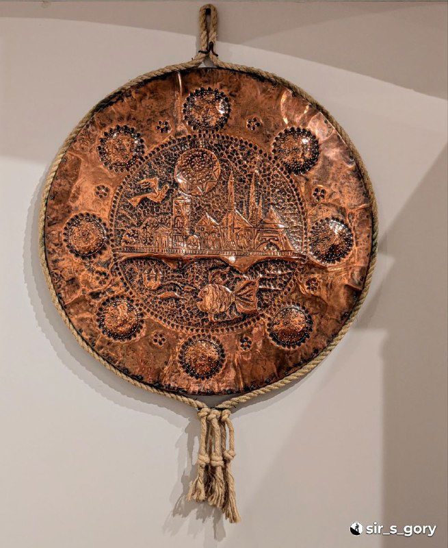Круглый медный предмет с чеканным орнаментом и сценой в центре — либо щит, либо поднос, представленный как образец прикладного искусства в музее.