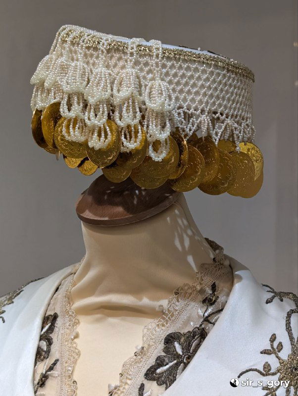 Женский головной убор с пришитыми монетами и кружевной отделкой на манекене — элемент традиционного костюма, демонстрирующий семейный и статусный смысл.