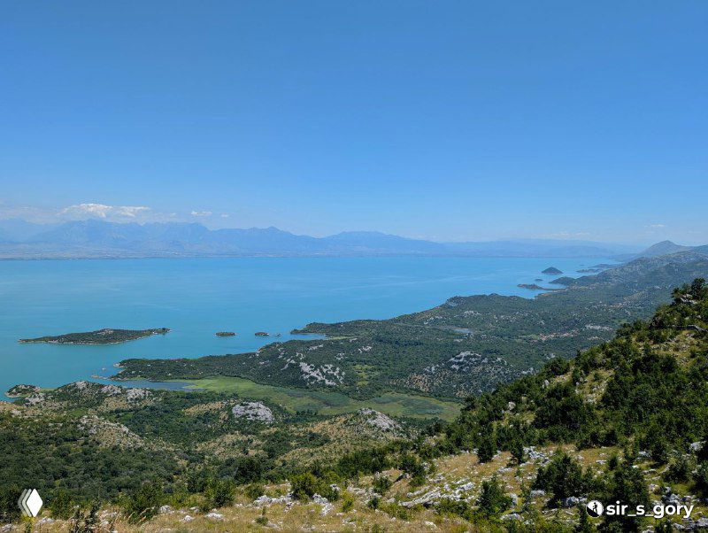 Широкая панорама бирюзовой глади Скадарского озера с островками и прибрежными склонами, переход к холмистой местности.