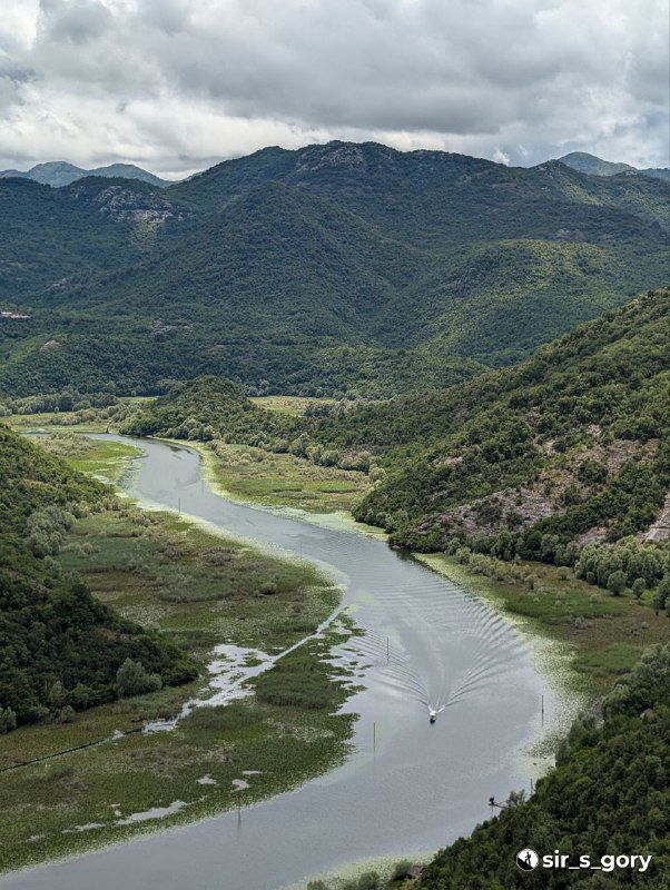 Река, втекающая в озеро, вид сверху с зелёных склонов; лодочная дорожка на воде и мелководные участки между островками.