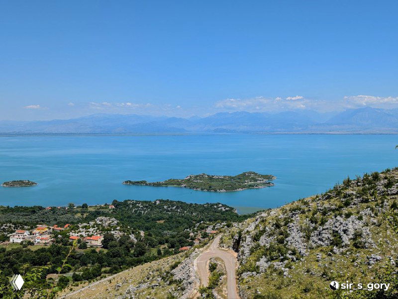 Вид с холма на водную гладь Скадарского озера, маленькие островки и прибрежные поселения, ясный день и дальние горы.