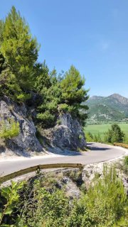 Извилистая серпантинная дорога у сосновых скал с видом на озёрные луга и холмы; летняя зелень и чистое синее небо.