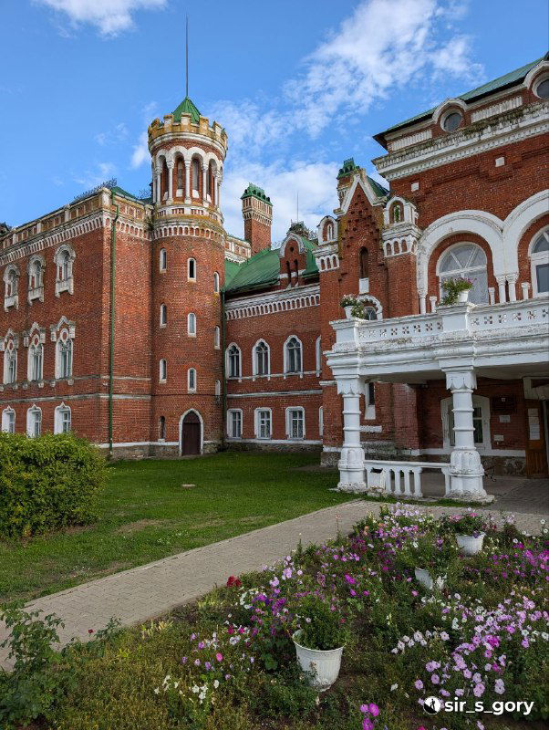 Крупный план угловой башни и цветочной клумбы возле входа усадьбы; кирпичная кладка, декоративные элементы и зелёные насаждения вокруг здания.