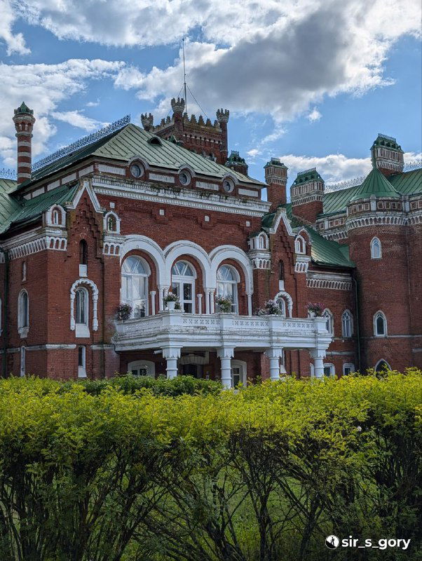 Парадный портик усадебного здания с арочными окнами и балконом, белые декоративные колонны на фоне красно-чёрной кирпичной фасадной отделки.