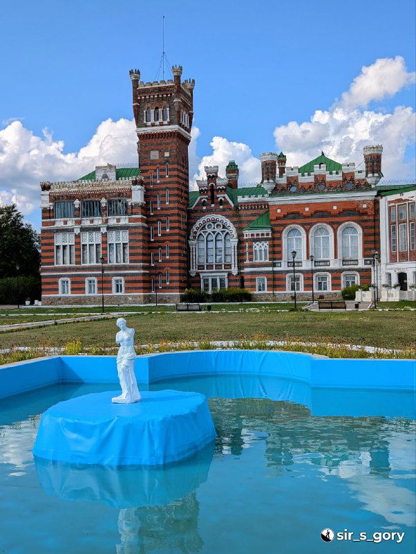 Фонтан на зелёном газоне перед усадьбой: голубая вода, скульптурный постамент и большая открытая лужайка, передний план композиции здания.