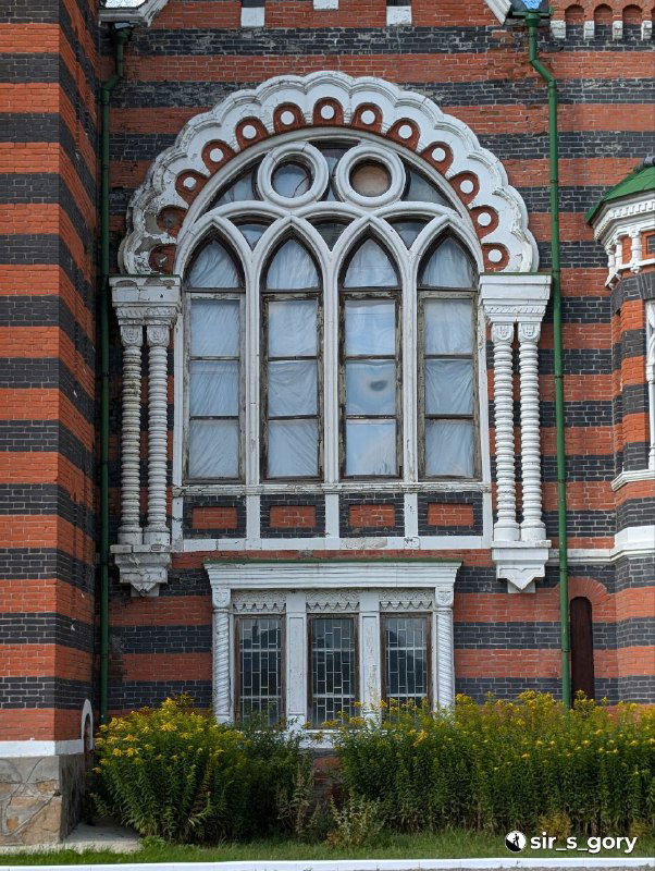 Деталь архитектурного окна в неоготическом стиле: арочное остекление, белый орнамент и красно-чёрная полосатая кирпичная кладка с растительностью у основания.