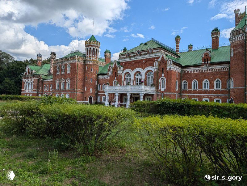 Боковой общий вид усадьбы сквозь зелёные кустарники: длинный фасад с башенками, арочными окнами и зелёными крышами под переменной облачностью.