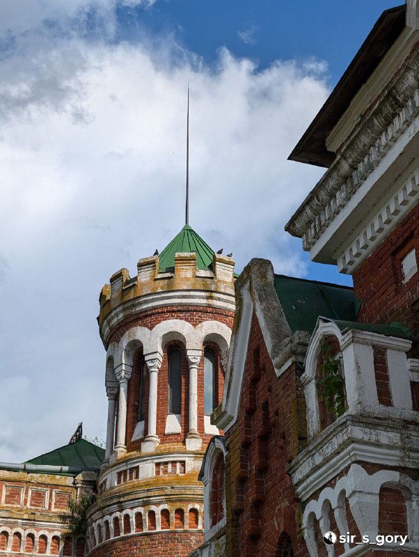 Крупная башенка с зубчатым верхом и узкими арочными окнами на фоне голубого неба; декоративный фрагмент внешней облицовки здания.