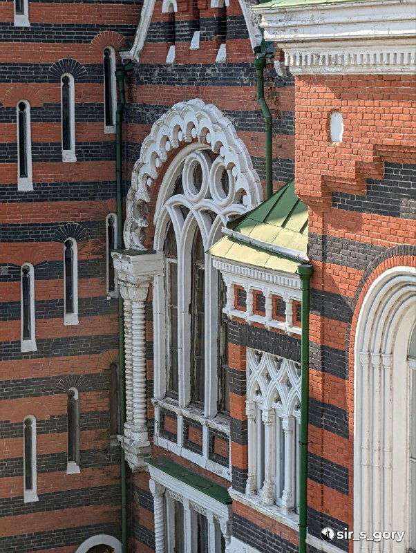 Фрагмент фасада с высокими арочными окнами и декоративными наличниками, вид под небольшим углом, растительность у фундамента и фактура кирпича.