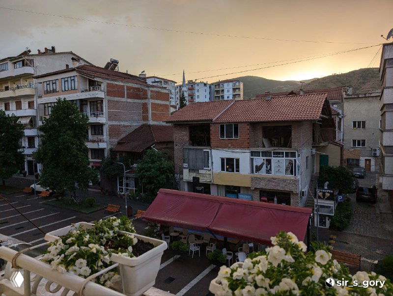 Вид с балкона отеля на соседние дома и улицу у озера в Поградеце, цветущие горшки на перилах и крыши на фоне холмов на закате.