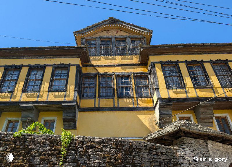Жёлтый фасад османского дома с деревянными эркерами и чёрными решётками, вид снизу через каменную подпорную стену, характерный городской фасад.