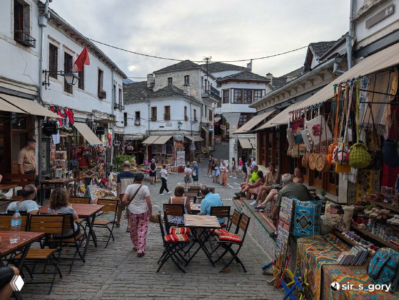 Узкая рыночная улица Гирокастры с туристами, торговыми лавками, уличными столиками и развешанными сувенирами, тесная атмосфера базара.