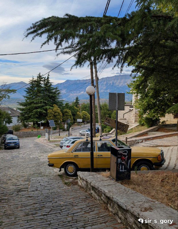 Жёлтый старый «Мерседес» припаркован у тротуара в Гирокастре, типичный городской автомобильный колорит на фоне горных видов и мощёных улиц.