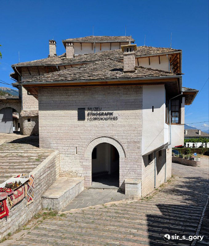 Фасад этнографического музея в доме-кула в Гирокастре с арочным входом, табличкой и мощёной дорожкой, здание в традиционном стиле 'куле'.