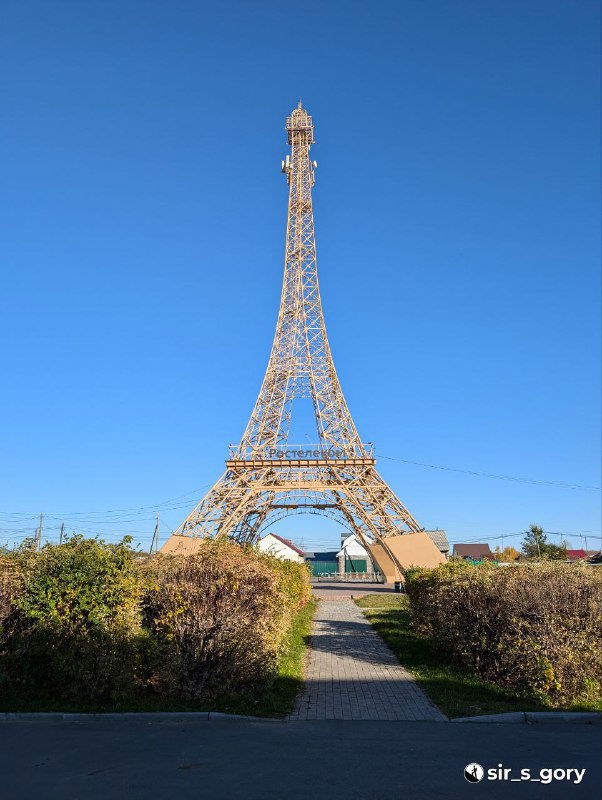 Вид спереди на местную копию Эйфелевой башни через декоративную зелёную стрижку и дорожку — точка притяжения для туристов в деревне.