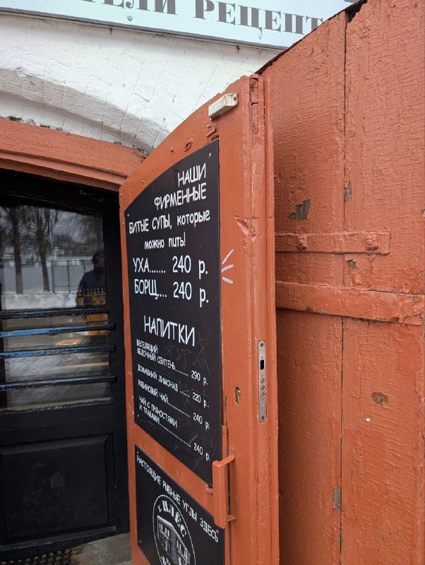 Уличная табличка с ценами на уху и напитки у входа в точку общепита, чёрная доска на фоне городского уличного пейзажа зимой.