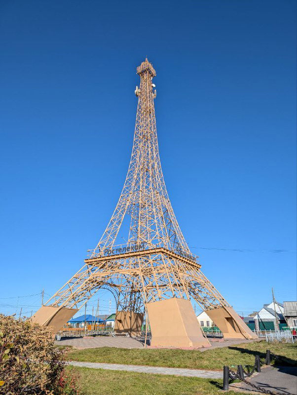 Высокая металлическая копия Эйфелевой башни в парковой зоне на фоне ясного неба; архитектурный объект и прогулочная аллея в дневном свете, фотография из серии.