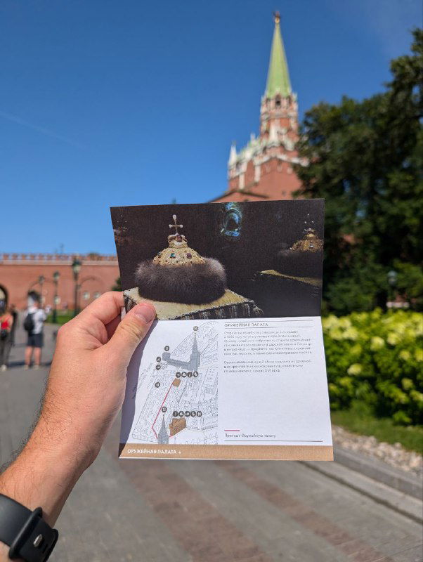 Рука держит открытку или путеводитель на фоне стен и башен Московского Кремля; туристический кадр с фокусом на бумажном материале и историческом фоне, дневное освещение.