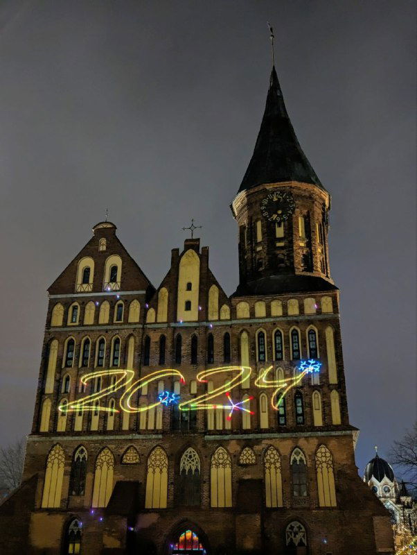 Ночной фасад старого кирпичного собора с подсветкой и световой инсталляцией «2024» на площади; праздничная иллюминация и архитектурный акцент в тёмное время суток.
