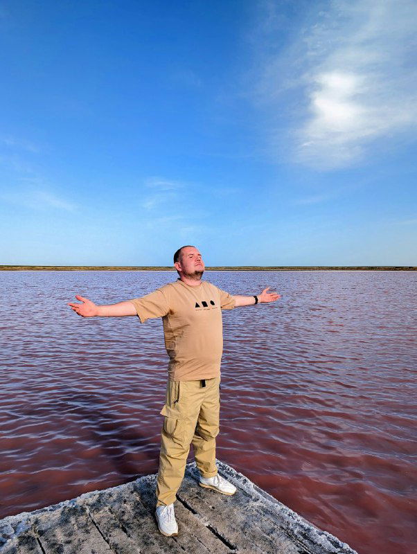 Человек стоит на берегу розового озера с разведёнными руками, простор плоской солёной поверхности и ясное небо; выразительный природный пейзаж, кадр автора.