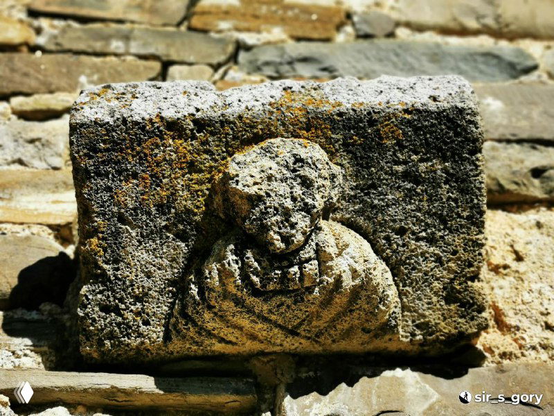 Малый декоративный каменный рельеф, напоминающий стилизованный лик или символ, крупный план фрагмента на стене Тхаба-Ерды.