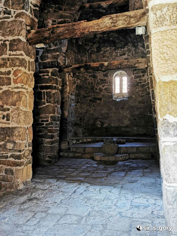 Интерьерный вид через низкую арку внутрь храма Тхаба-Ерды: каменные колонны, пол и небольшое узкое окно с мягким светом.