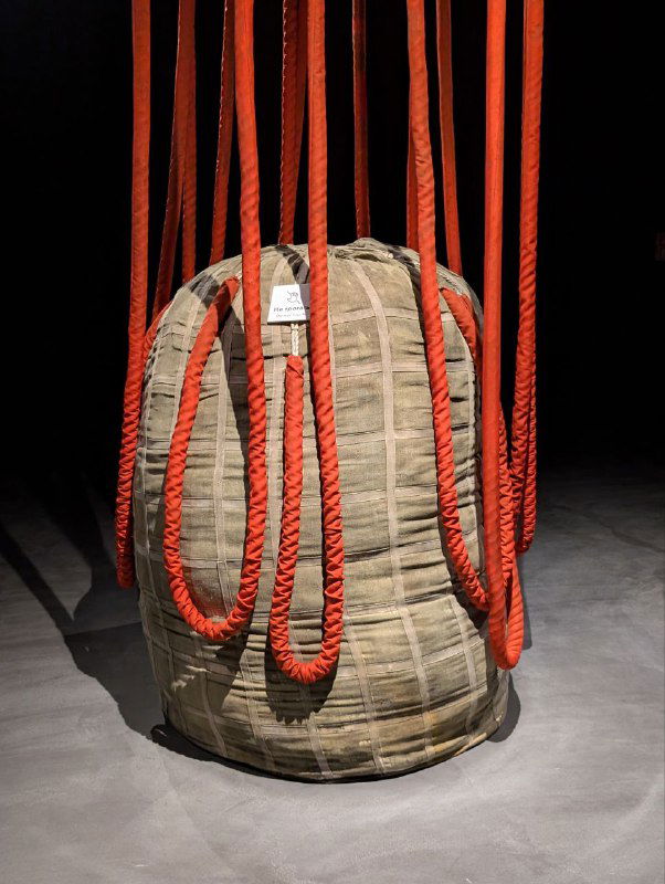 Инсталляция с подвешенным мешком и ярко-красными канатами в тёмном зале экспозиции музея «Атом», видно текстуру материалов и крепления.