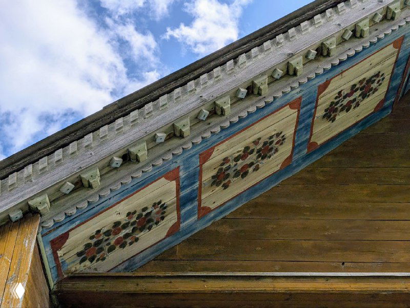 Декоративные раскрашенные панели под свесом крыши: квадратные поля с растительными мотивами на светлом фоне и резной карниз над ними